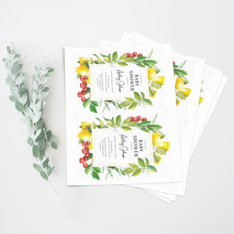 May include: A stack of baby shower invitations with a lemon and cherry design. The invitations are white with a gold border and feature the text "Baby Shower" and "Ashley Jones".
