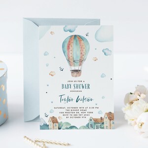 A watercolor baby shower invitation with a whimsical hot air balloon drifting over a cute village and green hills. The dreamy illustration includes soft pastel tones and playful details like puffy clouds and tiny rooftops. Customizable text appears in elegant fonts, ideal for a gender-neutral or adventure-themed baby shower.