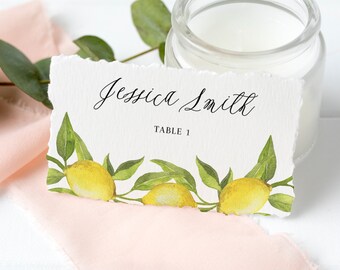 May include: A white place card with the name "Jessica Smith" in elegant script and "Table 1" in a smaller font. The card is decorated with watercolor lemons and green leaves. A lit candle in a glass jar and a pink ribbon are in the background.