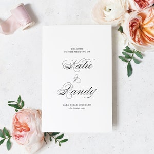 May include: A white wedding invitation with black calligraphy text. The text reads "Welcome to the wedding of Katie & Randy Lake Belle Vineyard 18th October 2021".