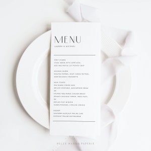 May include: A minimalist wedding menu card with a black and white design. The menu features a variety of dishes, including steak tartar, roasted peppers, pan seared striped bass, and hazelnut praline cake. The card is printed on high-quality paper and features a simple, elegant design.