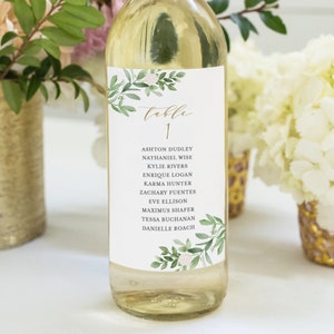 Könnte beinhalten: Eine Flasche Weißwein mit einem Etikett im grünen und weißen Blumendesign. Das Etikett trägt den Text "Tisch 1" und eine Namensliste: Ashton Dudley, Nathaniel Wise, Kylie Rivers, Enrique Logan, Karma Hunter, Zachary Fuentes, Eve Ellison, Maximus Shafer, Tessa Buchanan, Danielle Roach.