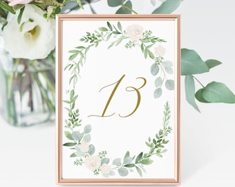 May include: A white card with a gold frame and a green floral wreath with white flowers. The number 13 is written in gold script in the center of the wreath.