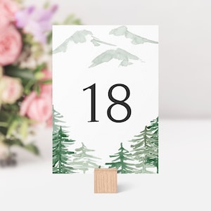 Op de afbeelding: Een aquarel tafelnummerkaart met een groene bos sc&egrave;ne en het nummer 18 in zwart gedrukt. De kaart is bevestigd aan een houten standaard.