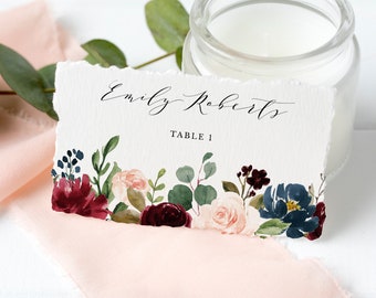 May include: A white wedding place card with a floral design in shades of burgundy, pink, and blue. The card reads "Emily Roberts" and "Table 1".