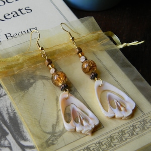 May include: A pair of gold earrings with brown and gold beads and a shell pendant. The earrings are hanging from a light brown fabric background.