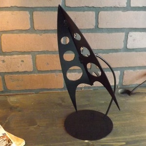 May include: A black metal sculpture of a stylized rocket ship with a round base. The rocket ship has a series of circular cutouts on the side.