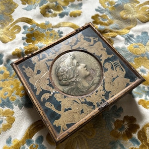 Puede incluir: Una caja de hojalata vintage con un borde dorado y un fondo negro. La caja presenta una imagen circular de una mujer con el pelo largo y una corona de flores. El texto "Biscuits Pernod" está impreso alrededor de la imagen.