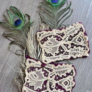 May include: Two decorative purple and cream lace pieces with a floral design are displayed with peacock feathers. The feathers have green and blue hues. Dried grass-like stems are also present.