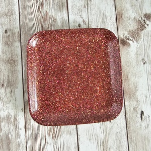 May include: A square, red glitter tray with a slightly raised edge. The tray is made of resin and has a sparkly, glittery finish.