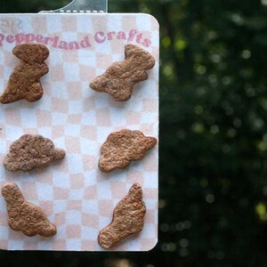 May include: A card with six brown, irregularly shaped objects, possibly cookies or ornaments, arranged on a pink and white checkered background. The card has the text "Pepperland Crafts" in pink script.