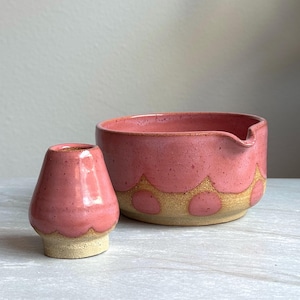 May include: A pink ceramic bowl and small vessel. The bowl has a scalloped design with a tan base and a spout. The vessel is a small, pink, cone-shaped object with a tan base. Both items have a speckled texture.
