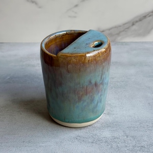 May include: A cylindrical ceramic cup with a glossy glaze featuring a blend of teal, blue, and brown tones. The top edge has a partial cover with a small circular opening, creating a unique silhouette for this handcrafted pottery vessel.
