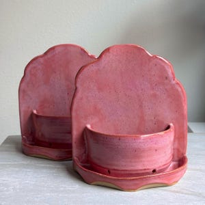 May include: Two handmade, pink ceramic wall planters. Each planter features a scalloped back, a small bowl, and a lower tray with drainage holes. The planters are glazed in a vibrant pink hue with subtle color variations.