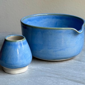 Puede incluir: Un juego de cuenco y chawan de matcha de cerámica azul. El cuenco tiene un pico y un borde verde. El chawan es un recipiente pequeño, azul y redondeado con una base blanca y un borde verde. El juego está diseñado para preparar té matcha.