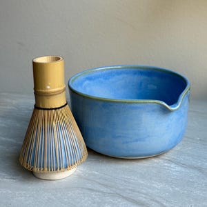 Może przedstawiać: Jasnoniebieska ceramiczna miska do matchy z dzióbkiem i bambusowa trzepaczka do matchy. Miska ma jasnozieloną obwódkę. Trzepaczka wykonana jest z bambusa i ma czarny sznurek wokół uchwytu.