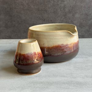 May include: A ceramic matcha whisk holder and a matching ceramic pouring bowl. Both pieces feature a two-tone glaze with a creamy off-white top section that transitions into a deep, rich reddish-brown base with a smooth, glossy finish.
