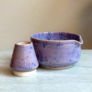 Puede incluir: Un juego de cuenco y soporte para batidor de té de cerámica. El cuenco es de color púrpura claro con un pico y el soporte para el batidor es un recipiente pequeño en forma de cono. Ambas piezas tienen un acabado esmaltado moteado con detalles en azul oscuro y una base blanca.