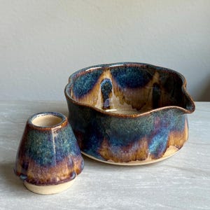 Puede incluir: Un juego de té de cerámica con una taza pequeña y redondeada y un cuenco más grande con un pico. Las piezas tienen un acabado esmaltado con una mezcla de tonos azules, marrones y dorados. El juego está diseñado para ceremonias de té.