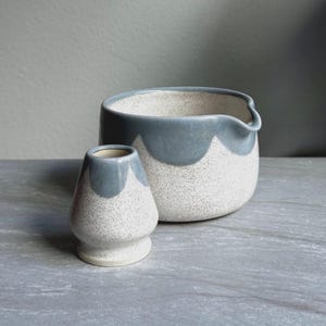 May include: A ceramic matcha bowl and a matching whisk holder featuring a speckled off-white base with a smooth, scalloped blue glaze along the upper rims. The bowl has a small pouring spout, and both pieces share a consistent speckled texture.