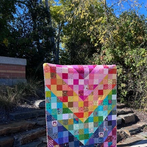 May include: A colorful patchwork quilt with a geometric design. The quilt is made up of squares of different colors and patterns, including pink, red, yellow, green, blue, and purple. The quilt is hanging on a clothesline in front of a brick wall and some greenery.