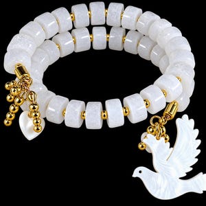 May include: A double-wrap bracelet featuring cylindrical white stone beads separated by small gold-toned metal spacers. The bracelet includes two dangling charms: a white heart-shaped bead and a white bird-shaped pendant, both attached with gold-toned metal links.