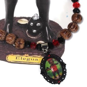 May include: A necklace featuring a pendant with a framed image of a figure in red clothing. The necklace includes wooden beads, red and black faceted beads, and a silver-tone clasp. The word "Elegua" is on a yellow label.