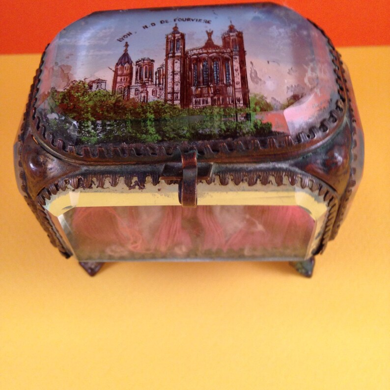 May include: An antique, rectangular trinket box with a hinged lid. The lid features a painted scene of a cathedral and the text "LYON - N.D. DE FOURVIERE". The box has a bronze-colored metal frame and glass sides, revealing pink contents.