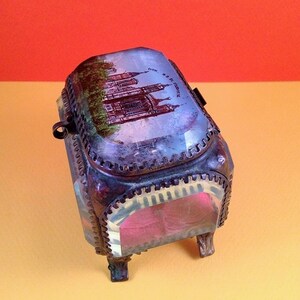 May include: Vintage metal trinket box with a hinged lid. The box features a detailed image of a building on the lid and sides. The box has a decorative trim and small feet. The interior is lined with pink fabric.