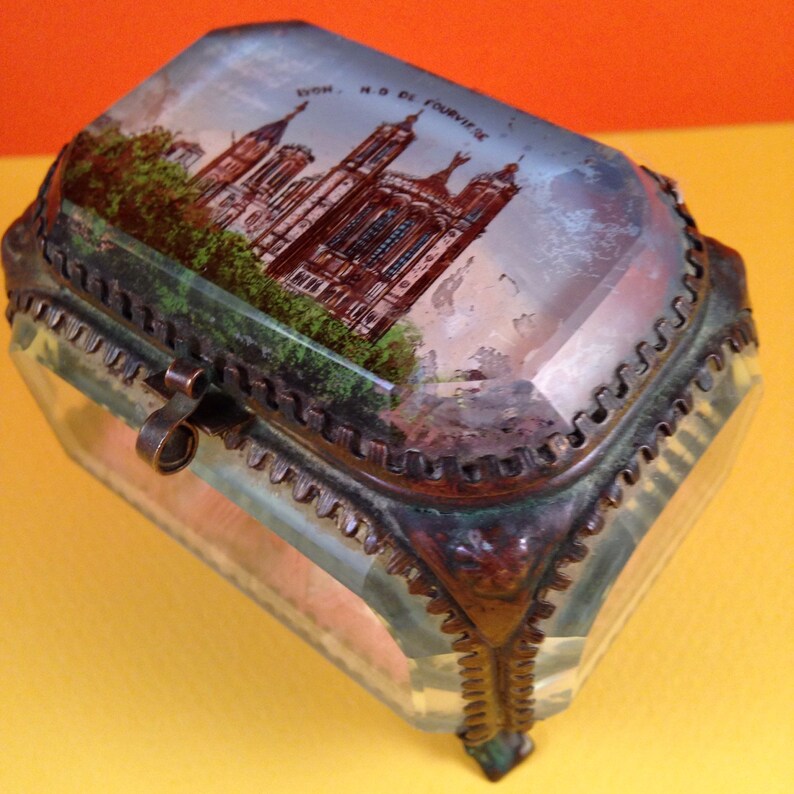 May include: A rectangular glass trinket box with a hinged lid. The lid features a painted image of a cathedral with the text "LYON, N.D. DE FOURVIERE". The box has a metal frame and clasp.