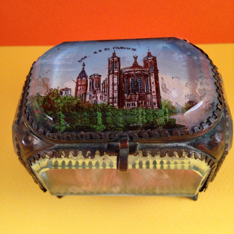 May include: An antique, rectangular trinket box with a hinged lid. The lid features a painted scene of a cathedral with the text "LYON, N.D. DE FOURVIERE". The box has a metal frame and a glass base.