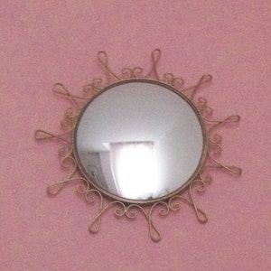 Puede incluir: Un espejo redondo con un marco dorado en forma de sol. El espejo est&aacute; montado en una pared rosa. El marco tiene remolinos y bucles decorativos que se extienden hacia afuera desde el espejo circular.