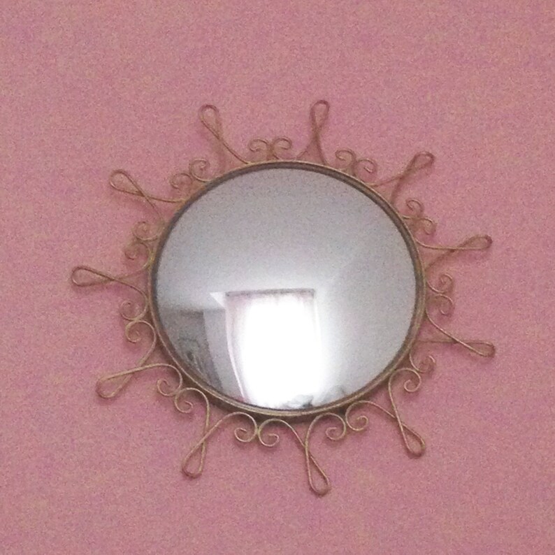 Puede incluir: Un espejo redondo con un marco dorado en forma de sol. El espejo est&aacute; montado en una pared rosa. El marco tiene remolinos y bucles decorativos que se extienden hacia afuera desde el espejo circular.
