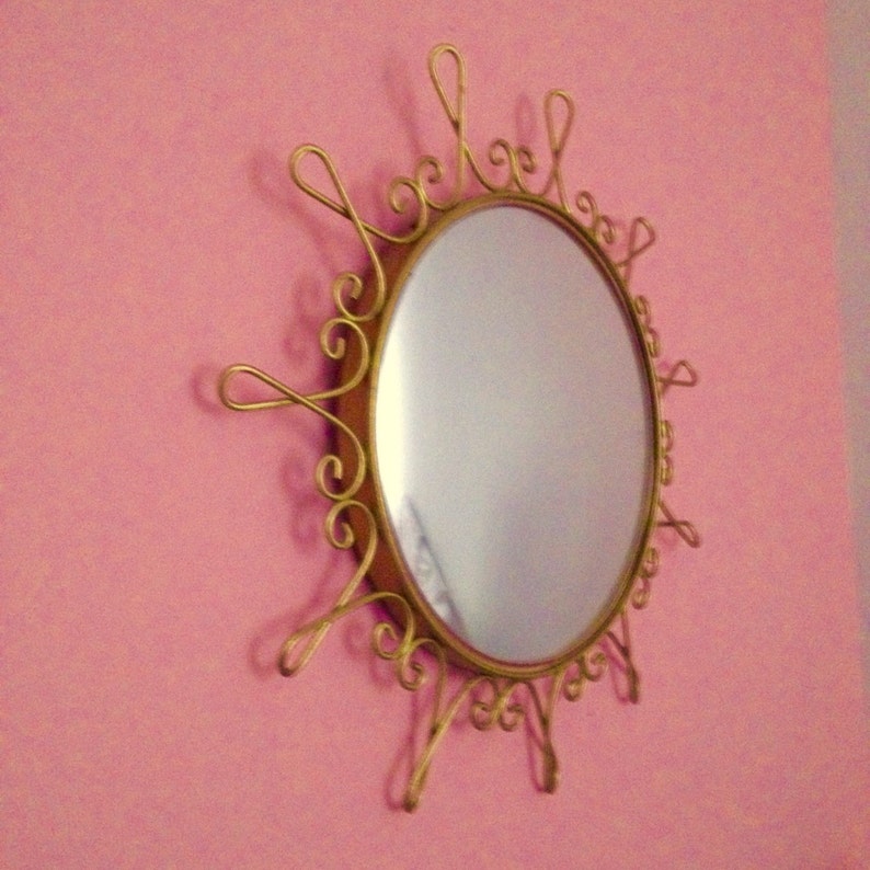 Puede incluir: Espejo ovalado con un marco dorado en forma de sol. El espejo est&aacute; montado en una pared rosa. El marco tiene detalles decorativos y ondulados alrededor del borde del espejo.