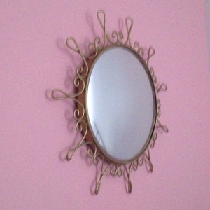 Puede incluir: Espejo de pared decorativo con marco de metal dorado con dise&ntilde;o de sol. El espejo tiene forma ovalada y est&aacute; montado en una pared rosa. El marco presenta detalles ornamentados y arremolinados.