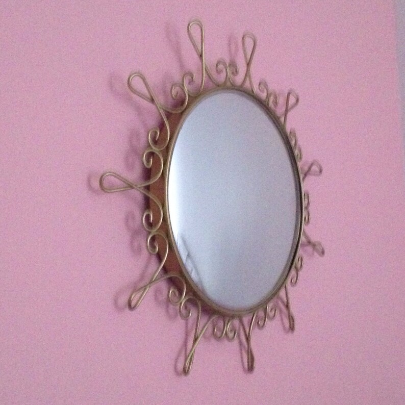 Puede incluir: Espejo de pared decorativo con marco de metal dorado con dise&ntilde;o de sol. El espejo tiene forma ovalada y est&aacute; montado en una pared rosa. El marco presenta detalles ornamentados y arremolinados.