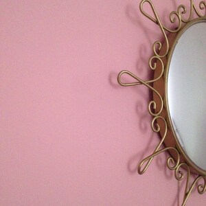 Puede incluir: Espejo ovalado de estilo vintage con un marco de metal dorado ornamentado. El marco presenta un dise&ntilde;o de sol con espirales y bucles decorativos. El espejo est&aacute; montado en una pared rosa, creando una est&eacute;tica retro.