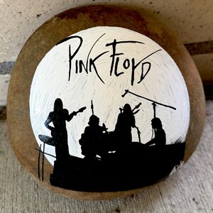 Könnte beinhalten: Ein handbemalter Stein mit dem Pink Floyd-Design. Der Stein zeigt den Namen der Band in schwarzer Schrift über einem weißen Kreis, mit einer schwarzen Silhouette der Band beim Auftritt. Der Stein hat eine natürliche, erdfarbene braune Farbe.