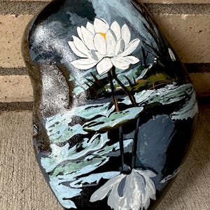 Könnte beinhalten: Ein glatter, dunkler, ovaler Stein, der von Hand mit einer weißen Seerose bemalt wurde. Das Kunstwerk zeigt eine zentrale weiße Blüte mit gelber Mitte, grüne Seerosenblätter und eine Spiegelung der Blume im dunklen Wasser darunter.