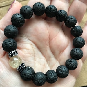 Könnte beinhalten: Schwarzes Lava-Stein-Armband mit einer klaren Quarzkristallperle und silbernen Akzenten.