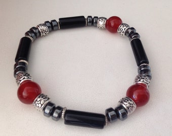 May include: A beaded bracelet featuring a combination of black, red, and silver-toned beads. The bracelet includes cylindrical black onyx beads, round red beads, and silver-toned decorative beads, strung together with smaller, dark gray beads.