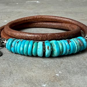 Puede incluir: Una pulsera envolvente con un cordón de cuero marrón y una fila de cuentas turquesas. La pulsera tiene un cierre plateado y un pequeño dije ovalado. El cordón de cuero está enrollado sobre sí mismo.