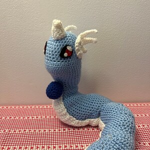 May include: A crocheted blue and white plush toy shaped like a dragon with red eyes. The dragon has a white horn and white spikes on its head. The dragon is curled up on a red and white checkered surface.