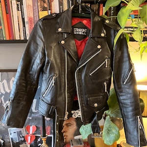 May include: Black leather motorcycle jacket with a zipper closure and silver hardware. The jacket has a red lining with a patch that says "Straight to Hell".