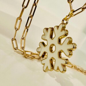 May include: A gold-toned necklace featuring a paperclip-style chain and a central snowflake pendant. The snowflake pendant is finished with a smooth, white enamel inlay, creating a contrast against the metallic gold edges of the jewelry piece.