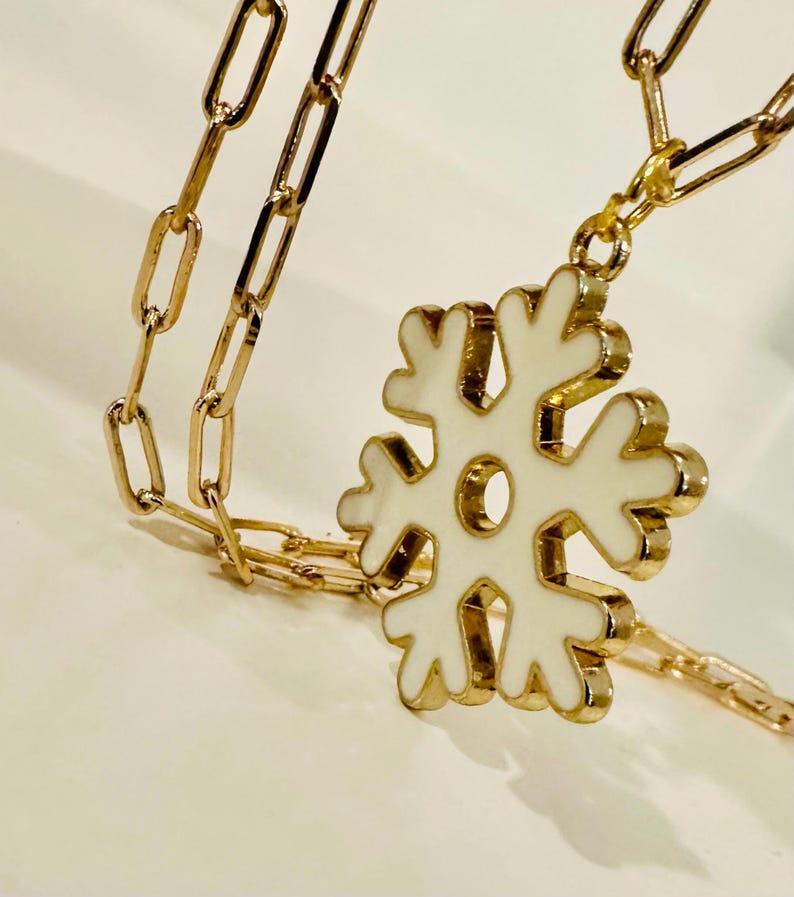 May include: A gold-toned necklace featuring a paperclip-style chain and a central snowflake pendant. The snowflake pendant is finished with a smooth, white enamel inlay, creating a contrast against the metallic gold edges of the jewelry piece.