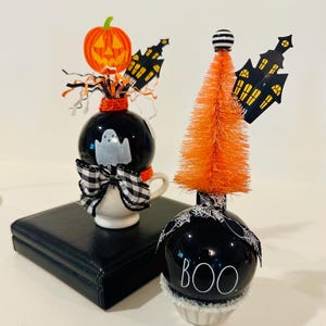May include: Two decorative Halloween ornaments featuring black spheres. One sits in a small white cup with a ghost, a pumpkin, and a plaid bow. The other is topped with an orange bottle brush tree, a black haunted house cutout, and a spiderweb ribbon with the text BOO.