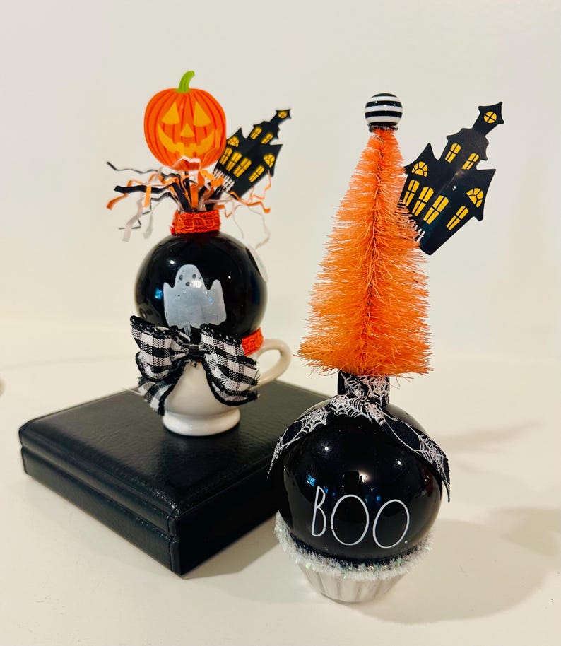 May include: Two decorative Halloween ornaments featuring black spheres. One sits in a small white cup with a ghost, a pumpkin, and a plaid bow. The other is topped with an orange bottle brush tree, a black haunted house cutout, and a spiderweb ribbon with the text BOO.