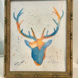 May include: A watercolor painting of a deer head with antlers in shades of blue and orange, featuring paint splatters on a white background. The artwork is enclosed in a rectangular gold frame with a textured, rope-like border design.