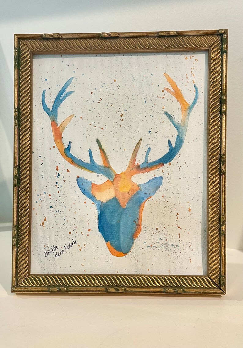 May include: A watercolor painting of a deer head with antlers in shades of blue and orange, featuring paint splatters on a white background. The artwork is enclosed in a rectangular gold frame with a textured, rope-like border design.